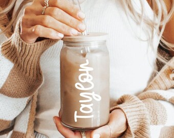 May include: A person holding a clear glass jar with a straw and the name "Taylor" written on it in white lettering.