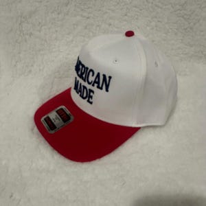 May include: A white and red baseball cap with the text "AMERICAN MADE" in navy blue. The cap has a red brim and a small red button on top. The cap is on a white, textured surface.