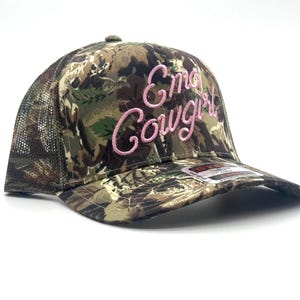 May include: Camouflage trucker hat with the text "Emo Cowgirl" embroidered in pink script. The hat features a mesh back and a curved brim. The front panel displays a woodland camouflage pattern.