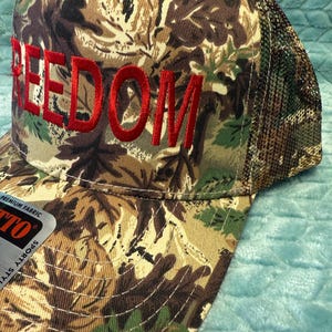 Puede incluir: Gorra de b&eacute;isbol de camuflaje con la palabra "FREEDOM" bordada en rojo. La gorra tiene una parte trasera de malla y una visera curva. La tela presenta un patr&oacute;n de marr&oacute;n, verde y beige.