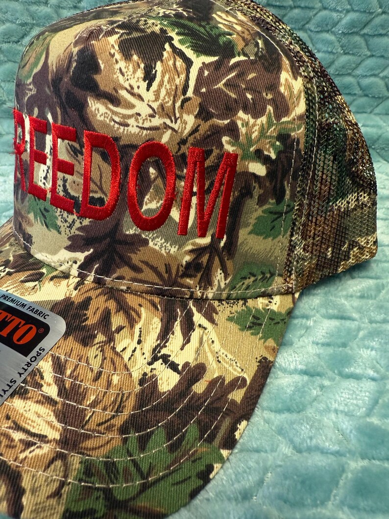 Puede incluir: Gorra de b&eacute;isbol de camuflaje con la palabra "FREEDOM" bordada en rojo. La gorra tiene una parte trasera de malla y una visera curva. La tela presenta un patr&oacute;n de marr&oacute;n, verde y beige.