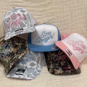 May include: A collection of trucker hats with the embroidered text "Emo Cowgirl". The hats feature various designs, including camouflage, a white and gray pattern, and solid colors. The text is in pink or blue.