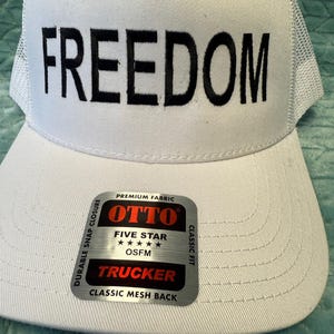 Puede incluir: Gorra de camionero blanca con la palabra "FREEDOM" en letras negras en negrita. La gorra tiene una parte trasera de malla y una visera curva. Una etiqueta en la visera dice "OTTO TRUCKER" y "CLASSIC MESH BACK".
