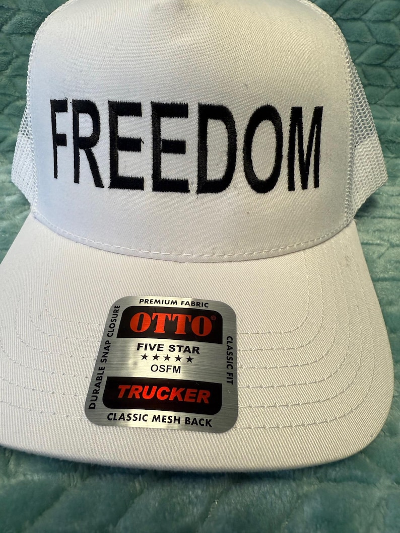 Puede incluir: Gorra de camionero blanca con la palabra "FREEDOM" en letras negras en negrita. La gorra tiene una parte trasera de malla y una visera curva. Una etiqueta en la visera dice "OTTO TRUCKER" y "CLASSIC MESH BACK".