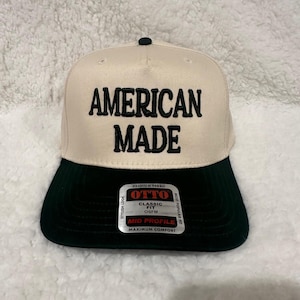May include: A beige and black baseball cap with the text "AMERICAN MADE" in bold black letters. The cap has a black brim and a small black button on top. The label reads "OTTO CLASSIC FIT OSFM MID PROFILE MAXIMUM COMFORT".