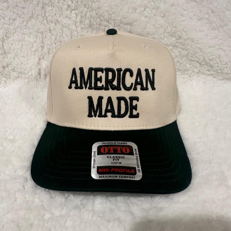 May include: A beige and black baseball cap with the text "AMERICAN MADE" in bold black letters. The cap has a black brim and a small black button on top. The label reads "OTTO CLASSIC FIT OSFM MID PROFILE MAXIMUM COMFORT".