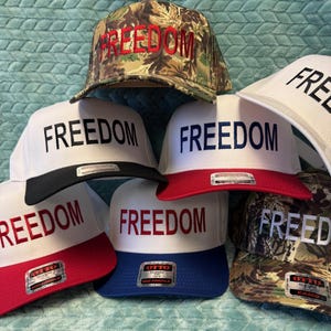 Puede incluir: Una colecci&oacute;n de gorras de b&eacute;isbol con la palabra "FREEDOM" bordada en la parte delantera. Las gorras presentan varias combinaciones de colores, incluyendo blanco con viseras rojas o negras, camuflaje y azul. El texto es rojo o negro.