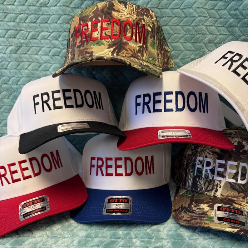Puede incluir: Una colecci&oacute;n de gorras de b&eacute;isbol con la palabra "FREEDOM" bordada en la parte delantera. Las gorras presentan varias combinaciones de colores, incluyendo blanco con viseras rojas o negras, camuflaje y azul. El texto es rojo o negro.