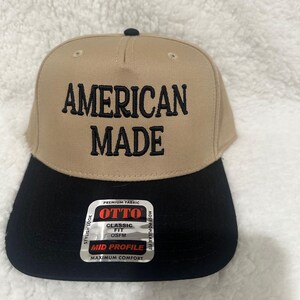 May include: A tan and black baseball cap with the words "AMERICAN MADE" embroidered in black. The cap has a black brim and a small black button on top. A sticker on the side reads "OTTO CLASSIC FIT OSFM MID PROFILE MAXIMUM COMFORT".