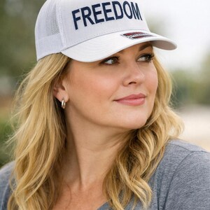 Puede incluir: Gorra de camionero blanca con la palabra "FREEDOM" bordada en azul marino en la parte delantera. La gorra tiene una parte trasera de malla y una visera curva. La persona lleva una camiseta gris.