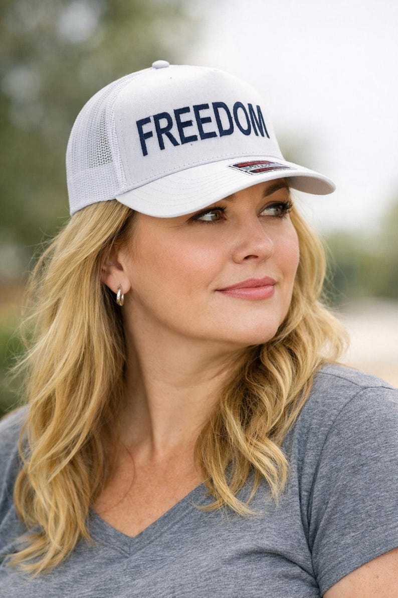 Puede incluir: Gorra de camionero blanca con la palabra "FREEDOM" bordada en azul marino en la parte delantera. La gorra tiene una parte trasera de malla y una visera curva. La persona lleva una camiseta gris.