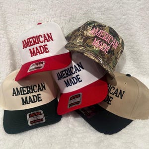 May include: A collection of baseball caps with the text "AMERICAN MADE" embroidered on the front. The caps come in various color combinations, including white and red, tan and black, and camouflage with pink lettering. Each cap has a flat brim.