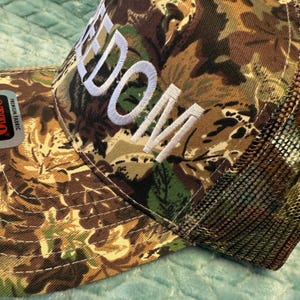Puede incluir: Gorra de camionero de camuflaje con la palabra "DOM" bordada en blanco. La gorra tiene una parte trasera de malla y una visera curva. El patr&oacute;n de camuflaje incluye tonos de marr&oacute;n, verde y beige.
