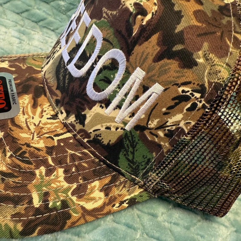 Puede incluir: Gorra de camionero de camuflaje con la palabra "DOM" bordada en blanco. La gorra tiene una parte trasera de malla y una visera curva. El patr&oacute;n de camuflaje incluye tonos de marr&oacute;n, verde y beige.