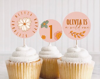 May include: Three pink and white cupcake toppers with floral designs. The toppers say "Olivia is a wild one", "Olivia Happy Birthday", and "1".
