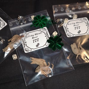 Könnte beinhalten: Drei durchsichtige Plastiktüten mit verschiedenen Glücksbringer und Kleinigkeiten. Jede Tüte hat ein weißes Etikett mit dem Text "Lucky Hand Root 777". Die Tüten enthalten auch grüne vierblättrige Kleeblätter, weiße Würfel und ein silbernes Hufeisen.