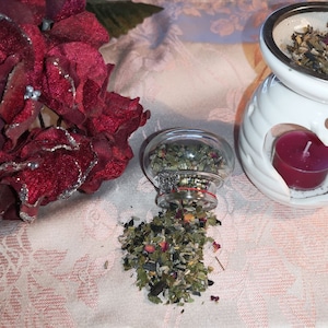 Peut inclure: Un diffuseur en céramique blanche avec une ouverture en forme de cœur et une bougie chauffe-plat à l'intérieur. Un pot en verre rempli d'herbes séchées se renverse sur une nappe en dentelle blanche. Une fleur de velours rouge est en arrière-plan.