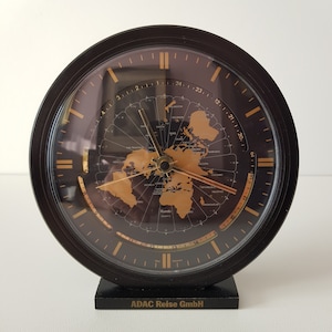 May include: A black world clock with gold accents and a map of the world on the face. The clock is on a black stand with the text "ADAC Reise GmbH" printed on the front.