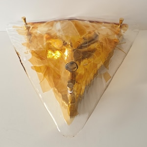 May include: A triangular wall sconce featuring a clear glass outer layer and an inner layer of amber-colored, textured glass shards. The fixture is secured with three small, metallic gold-toned hardware fasteners at the corners and center.