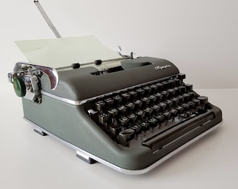 Op de afbeelding: Een groene vintage Olympia typemachine met een zwart toetsenbord en een zilveren retourwagenhendel. De typemachine heeft een papierhouder en een groene metalen behuizing.