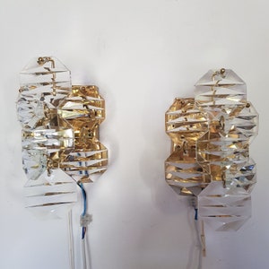 May include: Two gold-toned wall sconces with clear crystal geometric shapes. The sconces have a modern design and are perfect for adding a touch of elegance to any room.
