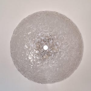 Könnte beinhalten: Eine Deckenleuchte aus klarem, strukturiertem Glas. Der runde Schirm hat eine blasenartige, unregelmäßige Oberfläche. Eine einzelne Glühbirne ist in der Mitte sichtbar. Die Leuchte ist an einer weißen Decke montiert.