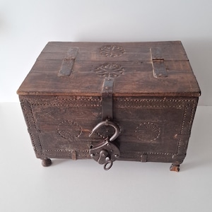 Peut inclure: Un coffre en bois brun foncé vieilli, orné de sculptures décoratives et de ferrures métalliques. Le coffre possède un couvercle à charnière, des sangles métalliques et un grand cadenas. Le bois a un aspect vieilli. Le coffre repose sur de petits pieds arrondis.