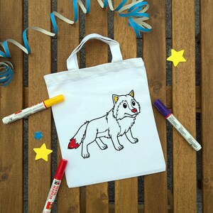 Könnte beinhalten: Eine weiße Stofftasche mit einer schwarz umrandeten Wolfsillustration, die gelbe Ohren, eine rote Nase und eine rot gefärbte Schwanzspitze aufweist. Die Tasche ist von gelben Sternen, blauen Luftschlangen und drei Textilmarkern in Rot, Gelb und Lila umgeben.