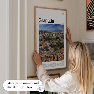 May include: A framed poster with the word "Granada" and a scenic view of the city. The poster is in a light wood frame and is being held up against a wall. The text "Mark your journey and the places you love" is at the bottom.