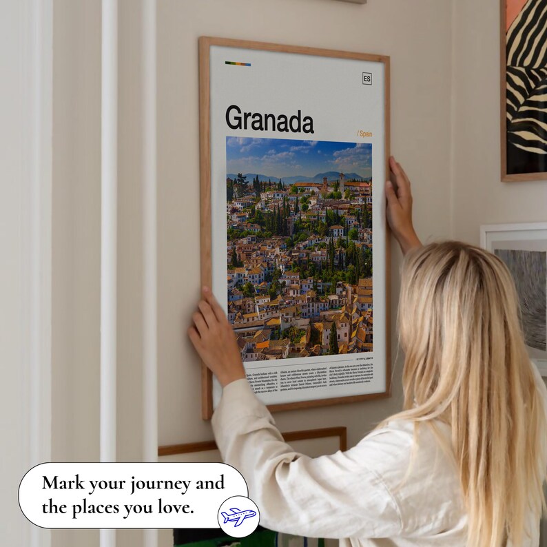 May include: A framed poster with the word "Granada" and a scenic view of the city. The poster is in a light wood frame and is being held up against a wall. The text "Mark your journey and the places you love" is at the bottom.