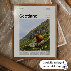 May include: A framed print featuring a Highland cow on a green hillside in Scotland. The print has the word "Scotland" in large text, with "United Kingdom" below. The print is packaged for safe delivery.