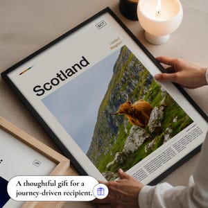 May include: Framed print featuring the word "Scotland" in large black font, with an image of a Highland cow on a green hillside. The print is in a black frame. Text at the bottom reads "A thoughtful gift for a journey-driven recipient."