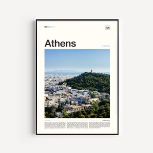May include: A framed print featuring a photograph of Athens, Greece. The image shows a cityscape with white buildings, a green hill, and a clear blue sky. The word "Athens" is printed in large, bold letters at the top.