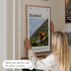 May include: A framed poster featuring a scenic view of Scotland, with the word "Scotland" in large text. The image shows a brown Highland cow on a green hillside. The poster is being held up on a wall.