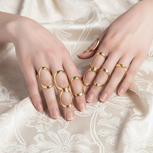 May include: Gold-colored finger rings in various designs, including full finger rings and simple bands. The rings are displayed on hands against a cream-colored, patterned background. The jewelry is elegant and eye-catching.