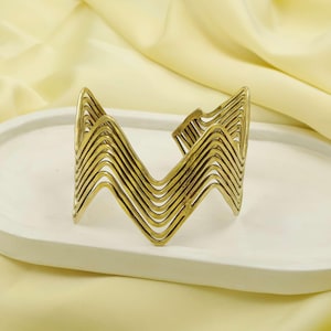 May include: A gold-coloured cuff bracelet with a layered, zigzag design. The bracelet is made of thin, curved metal strips that create a wave-like pattern. The jewellery is displayed on a white, oval-shaped tray.