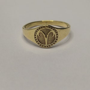 May include: A gold signet ring featuring a raised design of the astrological symbol for Aries, the ram.