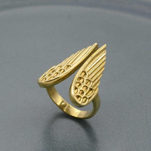 Puede incluir: Un anillo dorado con diseño de alas. El anillo presenta dos alas estilizadas que se curvan alrededor del dedo. Las alas tienen patrones de plumas detallados y una apariencia texturizada, creando un accesorio único y elegante.