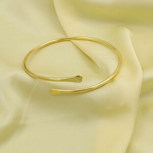 Könnte beinhalten: Ein goldfarbenes, offenes Armreif-Armband. Das Armband hat ein schlichtes, elegantes Design mit einem glatten, abgerundeten Band, das sich zu abgeflachten, leicht gebogenen Enden verjüngt. Der Armreif wird auf einer hellgelben Satin-Oberfläche präsentiert.