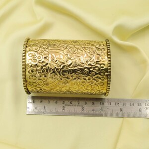 May include: A gold-colored, cylindrical container with intricate floral and leaf patterns. The container is approximately 4 inches long and has decorative beaded edges. The surface is highly reflective, and the container is set against a soft, yellow background.