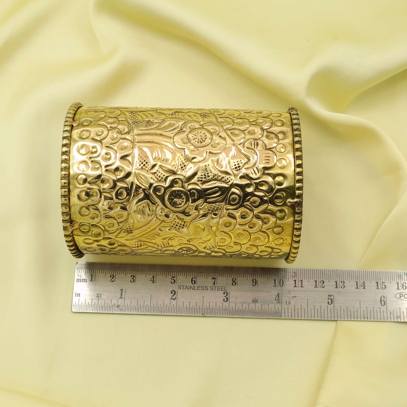 May include: A gold-colored, cylindrical container with intricate floral and leaf patterns. The container is approximately 4 inches long and has decorative beaded edges. The surface is highly reflective, and the container is set against a soft, yellow background.