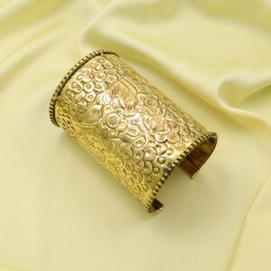 May include: A gold-colored cuff bracelet with an intricate floral pattern. The cuff is cylindrical with a textured surface and detailed design. The edges are lined with a beaded trim, and the bracelet is open on one side.
