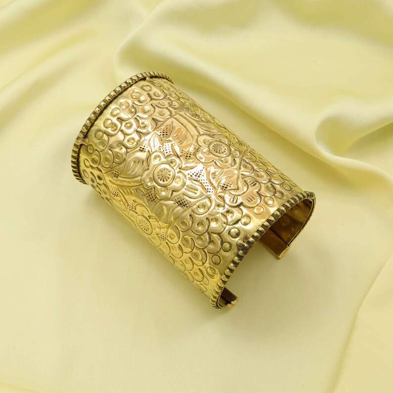 May include: A gold-colored cuff bracelet with an intricate floral pattern. The cuff is cylindrical with a textured surface and detailed design. The edges are lined with a beaded trim, and the bracelet is open on one side.