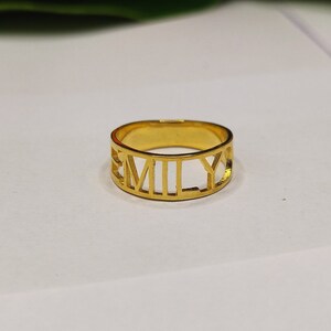 以下が含まれることがあります： 「EMILY」の文字が切り抜かれた、金色のリング。 リングはバンドスタイルで、文字が外側の表面を形成しています。 リングはおそらく金または金色の金属で作られています。