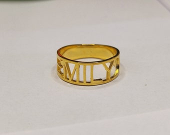 Op de afbeelding: Een goudkleurige ring met de naam "EMILY" uitgesneden in blokletters. De ring is een bandstijl, waarbij de letters het buitenoppervlak vormen. De ring is waarschijnlijk gemaakt van goud of een goudkleurig metaal.