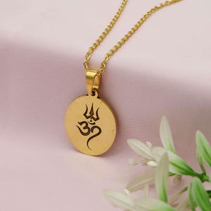May include: Gold-colored pendant necklace featuring a circular pendant with a black Om symbol and trident design. The necklace has a delicate chain and is displayed on a pink background with white flowers.