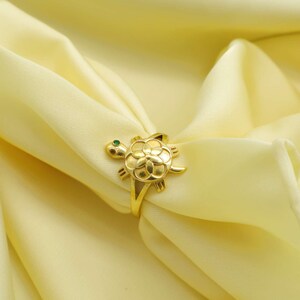 May include: A gold-colored ring in the shape of a turtle. The turtle has a flower-like design on its shell and a small green gem for an eye. The ring is set against a soft, yellow background.