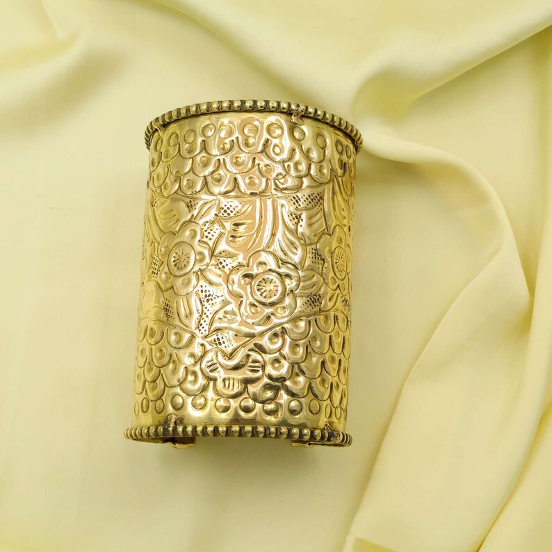 May include: A gold-colored cuff bracelet with intricate floral and leaf patterns. The cuff has a cylindrical shape with detailed embossed designs and a beaded border. The metal has a polished, shiny finish, reflecting light.