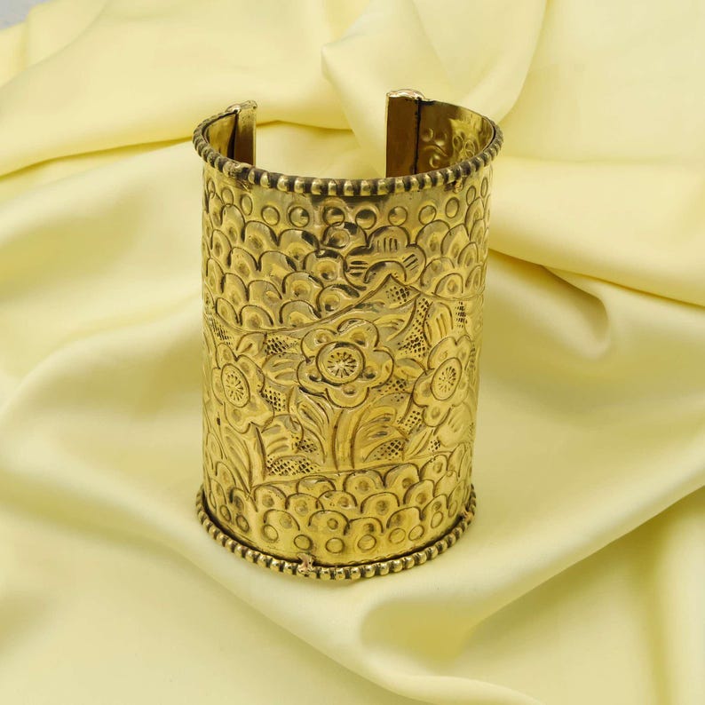 May include: Gold-colored cuff bracelet with an embossed floral design. The cuff is cylindrical with an open back and features detailed patterns of flowers and leaves. The edges are adorned with a beaded trim, creating a vintage aesthetic.