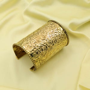 May include: A gold-colored cuff bracelet with an intricate floral pattern. The cuff is wide and wraps around the wrist, featuring detailed embossed flowers and leaves. The metal has a polished finish.